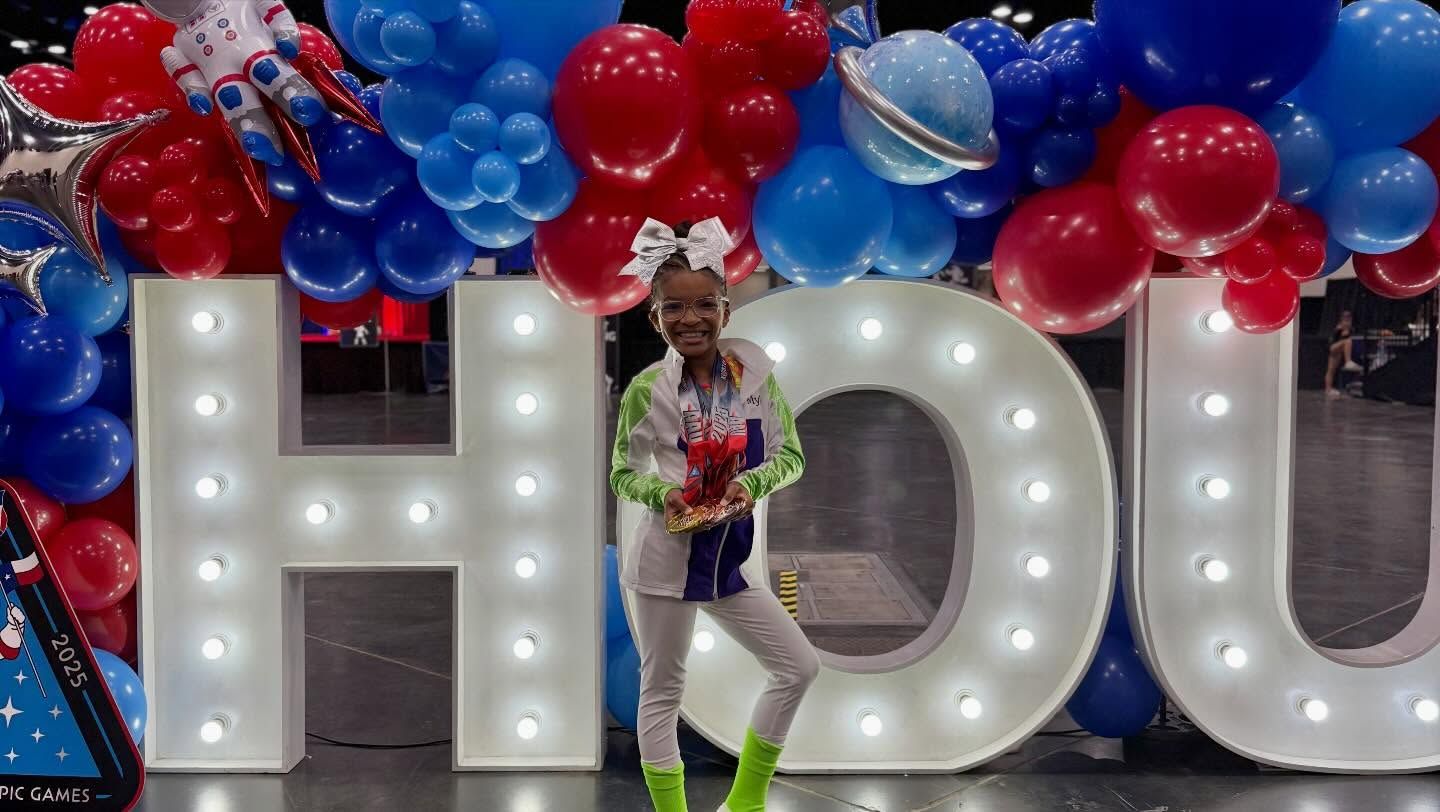 Person posing in front of lit “HOU” letters with red and blue balloons.