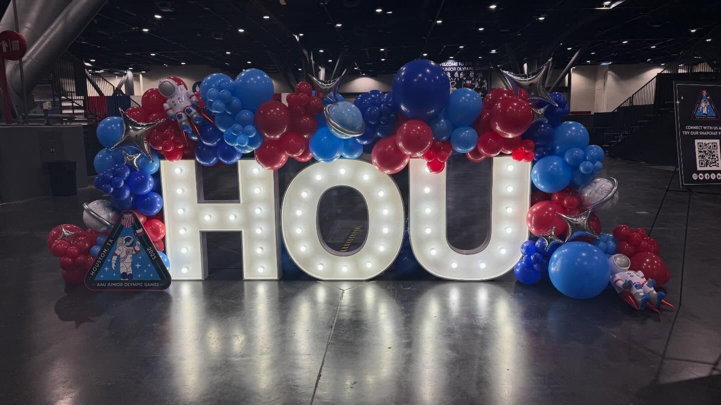 Illuminated “HOU” letters surrounded by red and blue balloons in a dark indoor venue