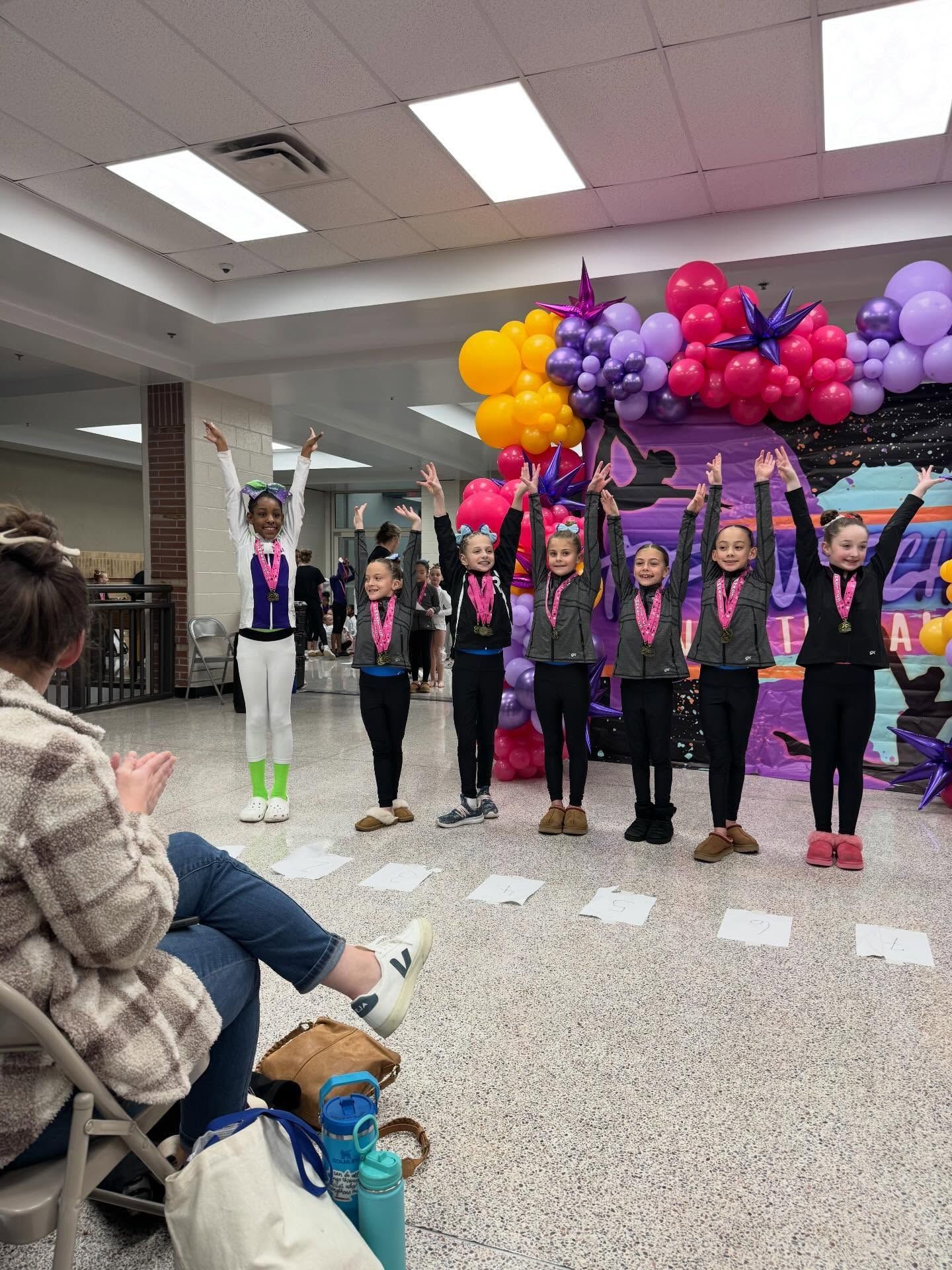 Children in matching outfits pose under colorful balloon arches in a community room
