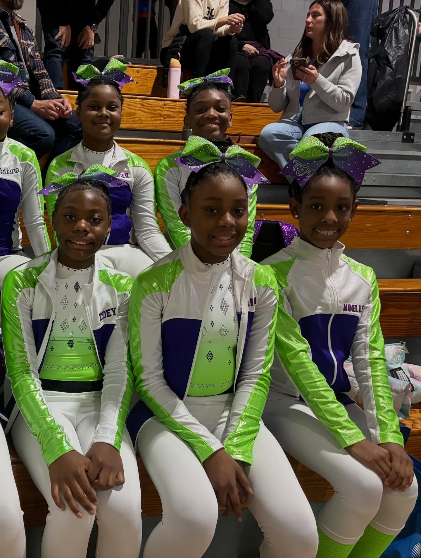 Five girls in green, white, and purple outfits sit in rows, smiling at a performance event.