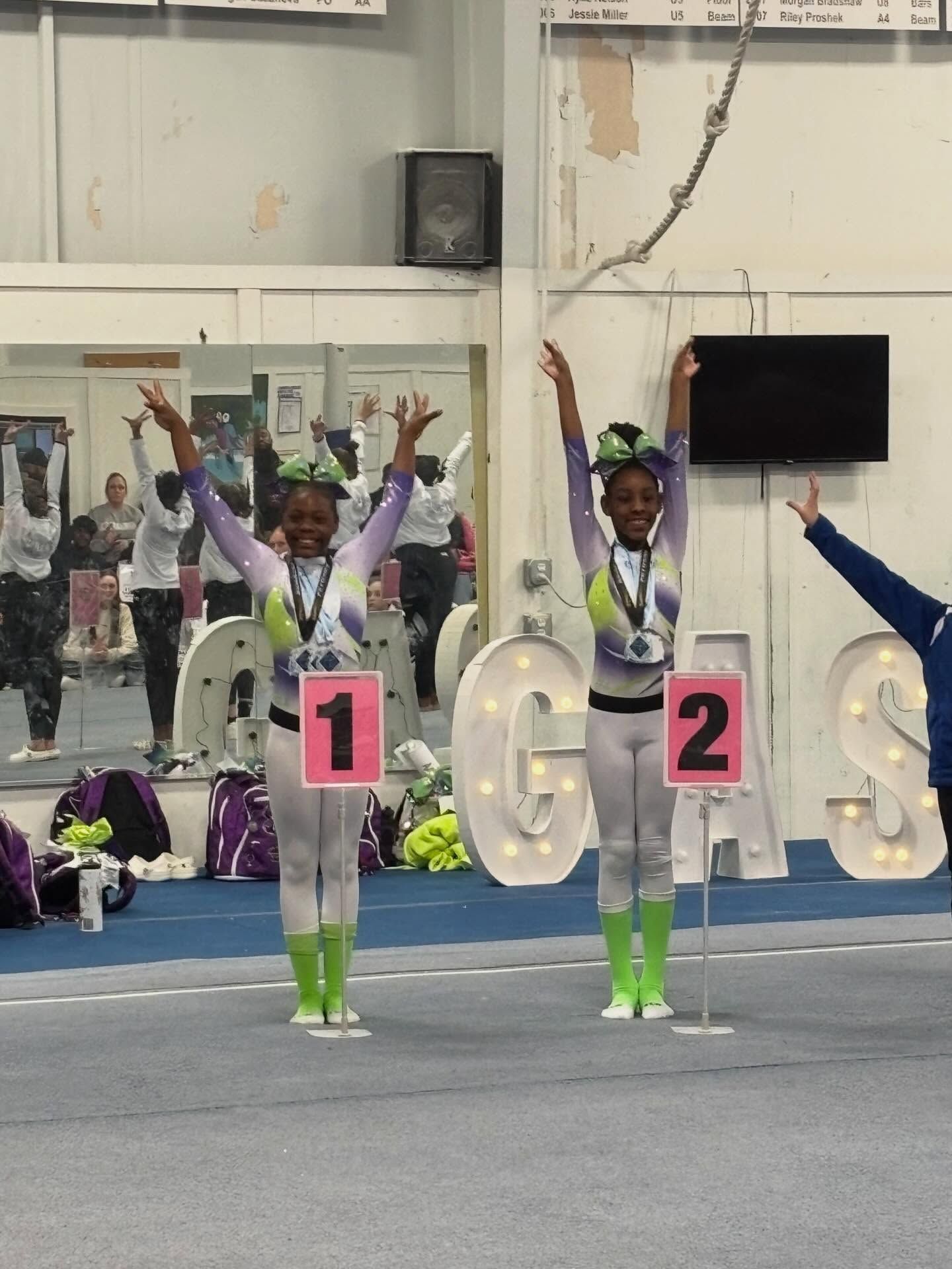 Two cheerleaders in purple and white uniforms pose with arms raised beside lit letters 