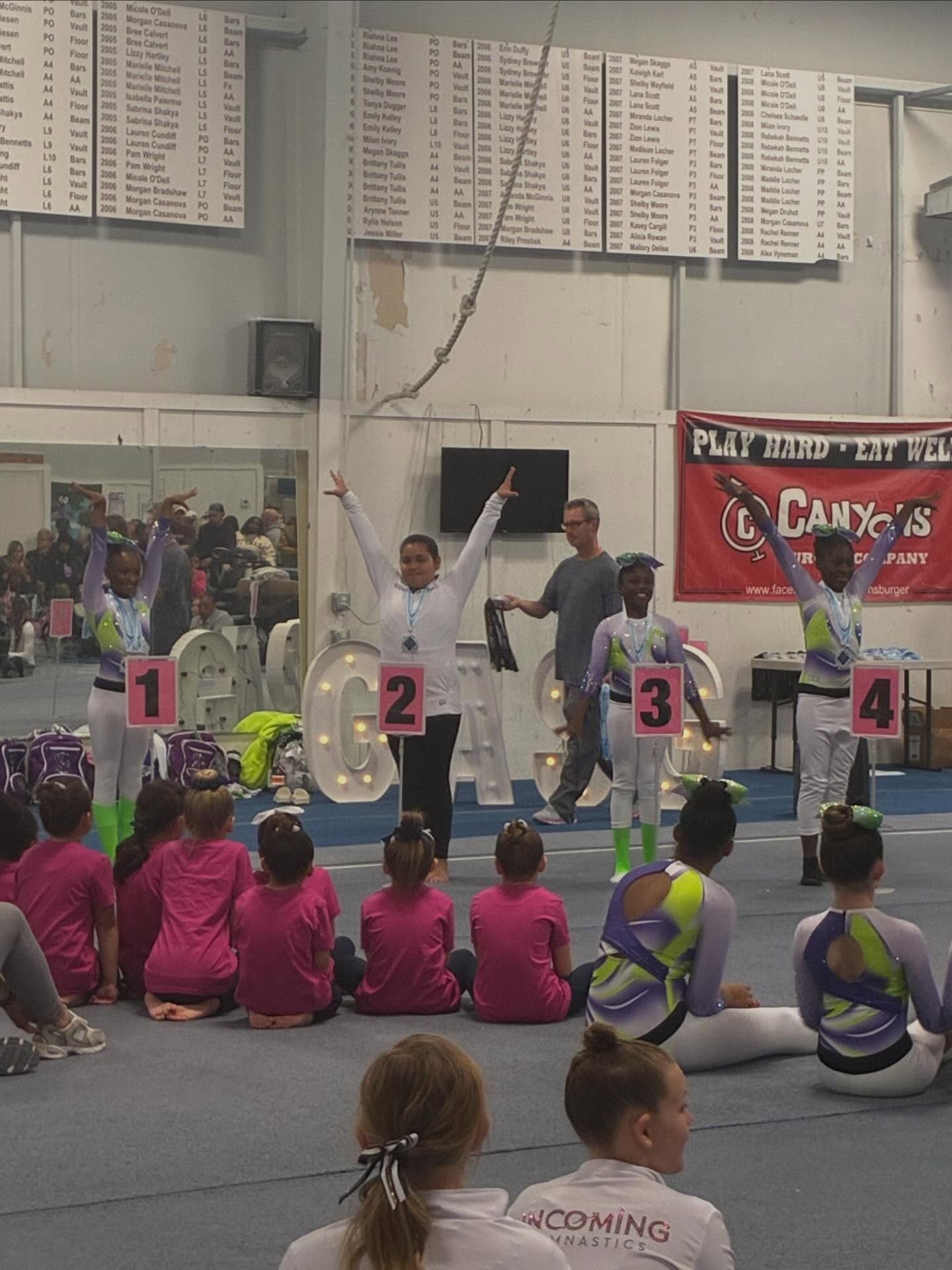 Indoor gym award ceremony with winners on podium numbers 1–4 and children seated in front
