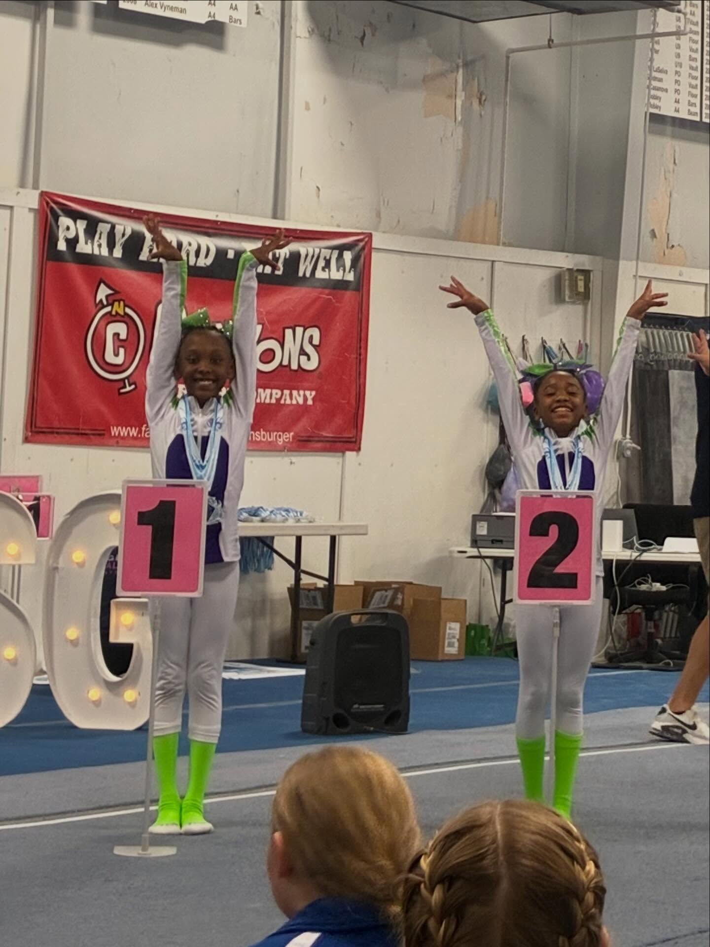 Two gymnasts on podiums labeled 1 and 2 with arms raised in a gymnasium.