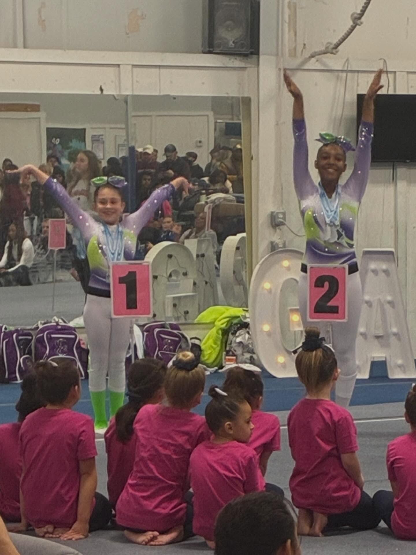 Two gymnasts pose on podiums labeled 1 and 2 while seated spectators watch in a gymnasium.