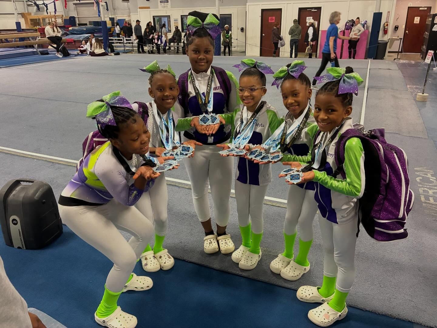 Five cheerleaders in purple and green uniforms pose together on a gym mat, smiling and holding pom-poms.
