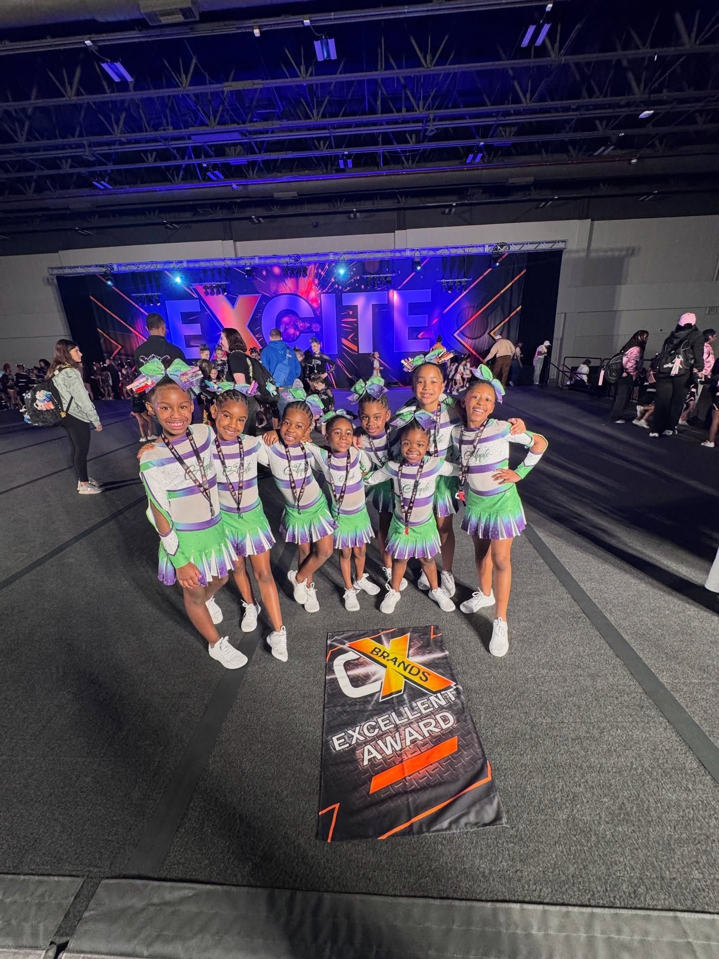 Cheer team posing on a competition floor in green, purple, and white uniforms with a lit stage backdrop