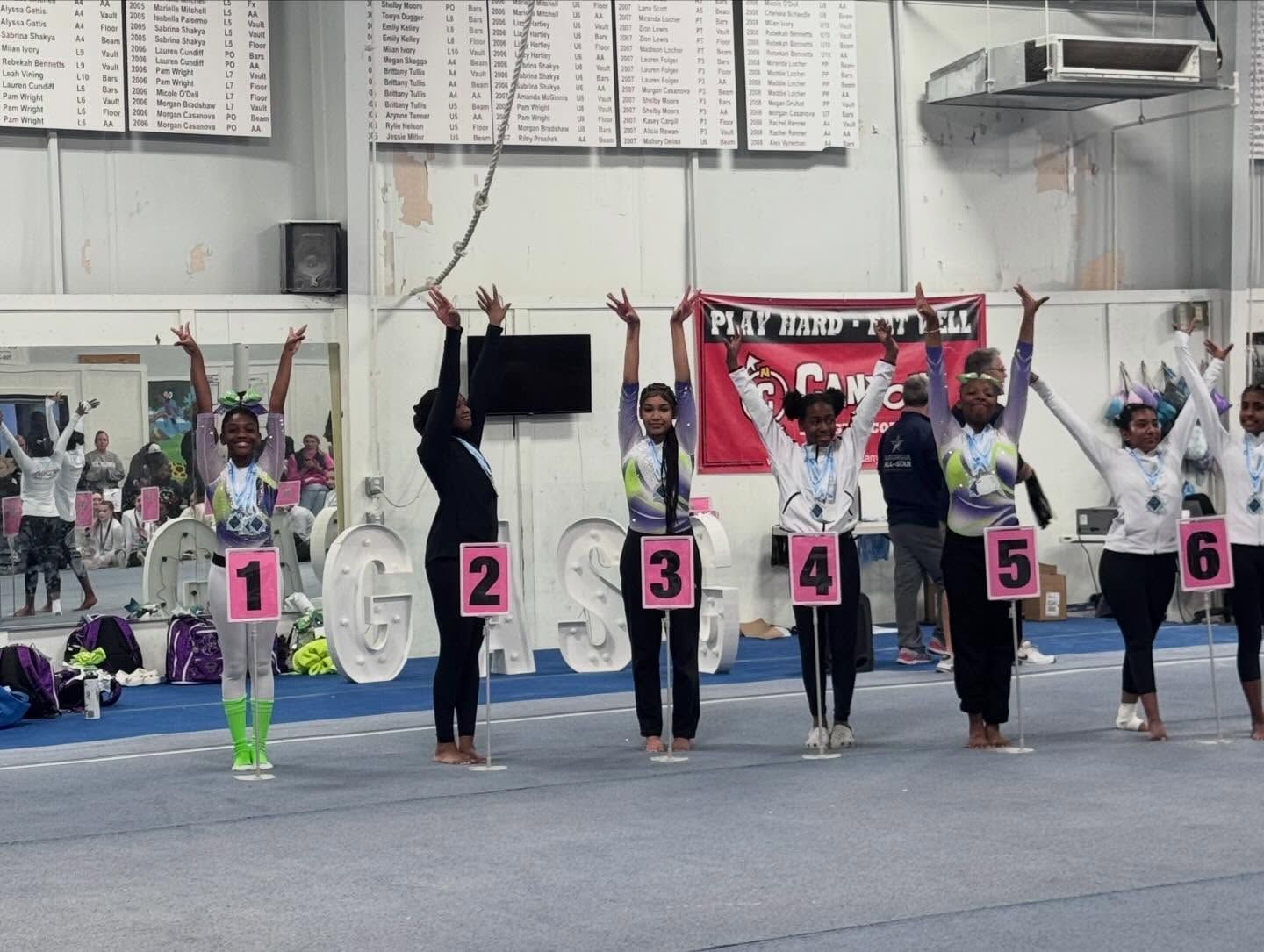 Six athletes stand with raised arms behind numbered markers in an indoor gym.