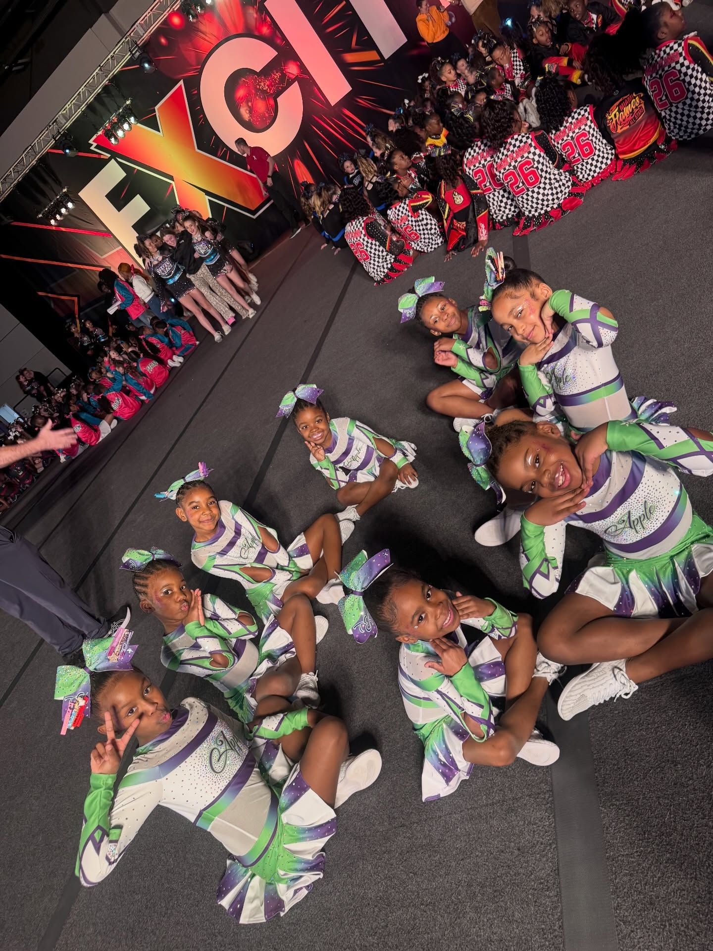 Cheer squad posing on a city street at a parade, wearing green-and-white uniforms with raised pom-poms.