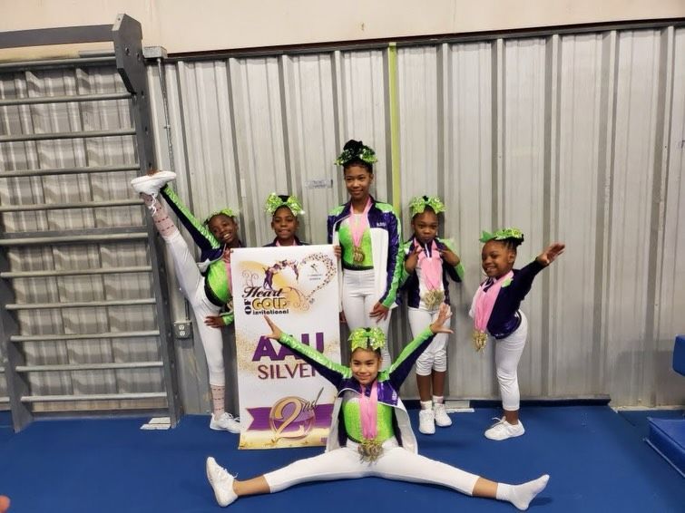 Young gymnasts in pink, green, and white pose with a silver award banner in a gymnasium