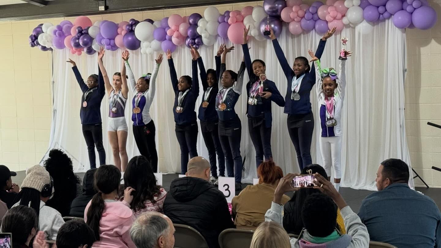 Children on a stage reaching up under purple and pink balloon decorations, with an audience watching.