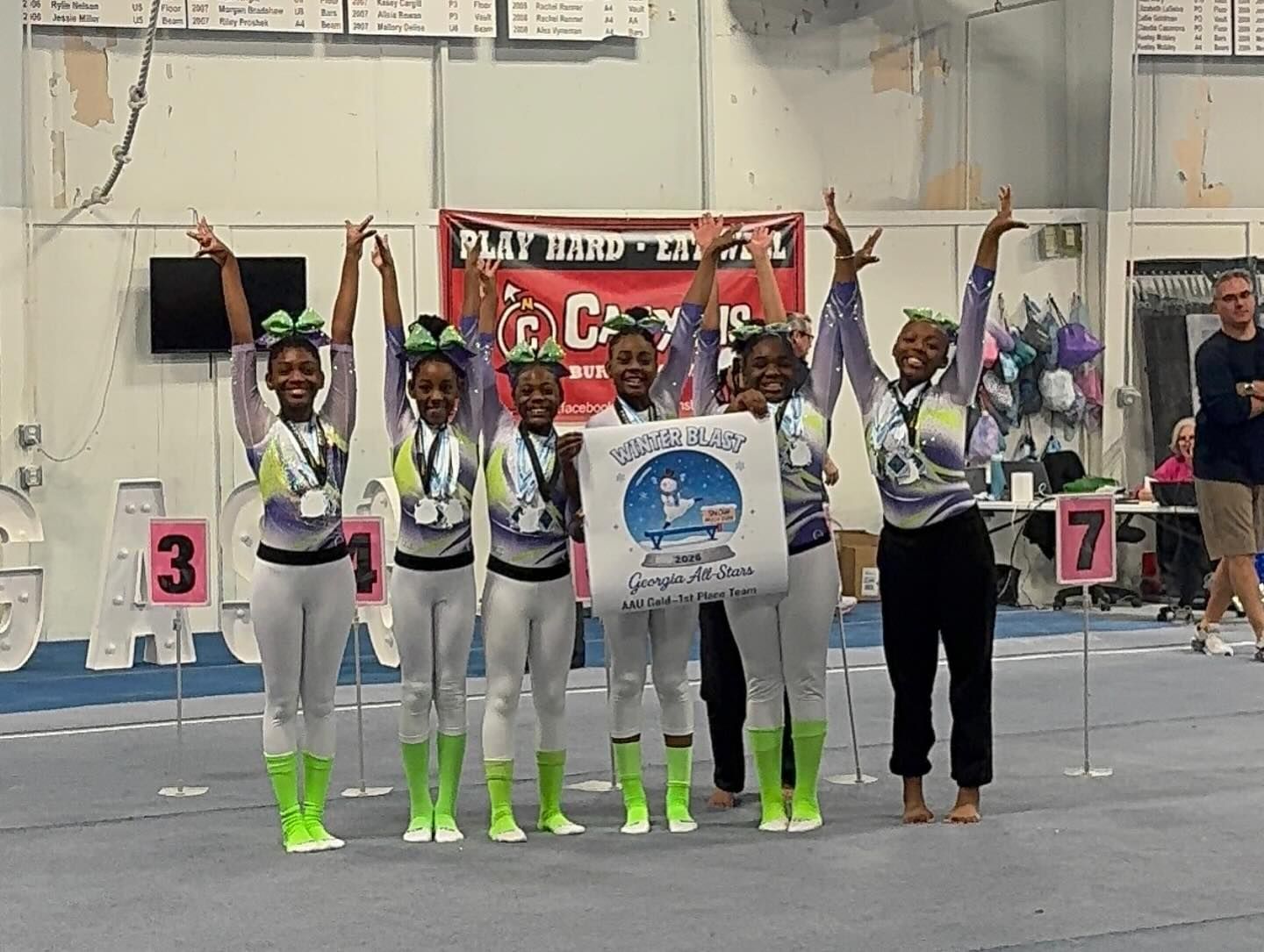 Five gymnasts in medals pose with raised arms at an indoor competition podium