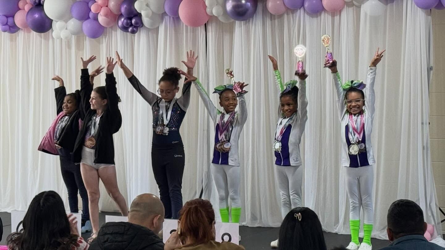 Five performers on stage raise their arms during a dance routine under purple and white balloons.