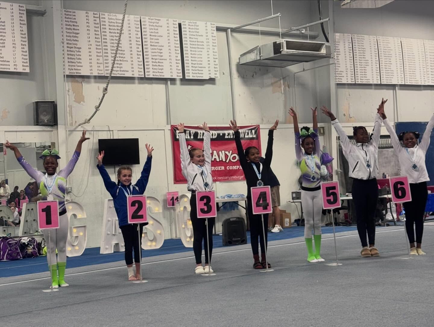 Gymnastics team lined up indoors with arms raised, wearing numbered bibs and bright leg warmers