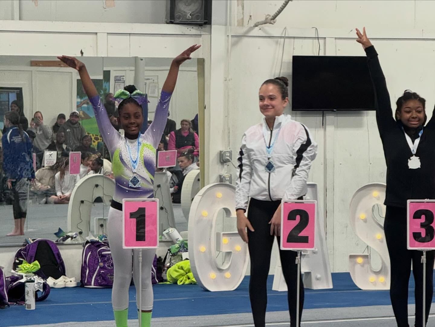 Three gymnasts on a podium with medals, arms raised in celebration, at an indoor competition.