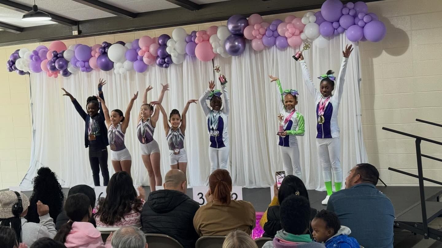 Children onstage beneath purple balloons, raising their arms during a performance in front of an audience.