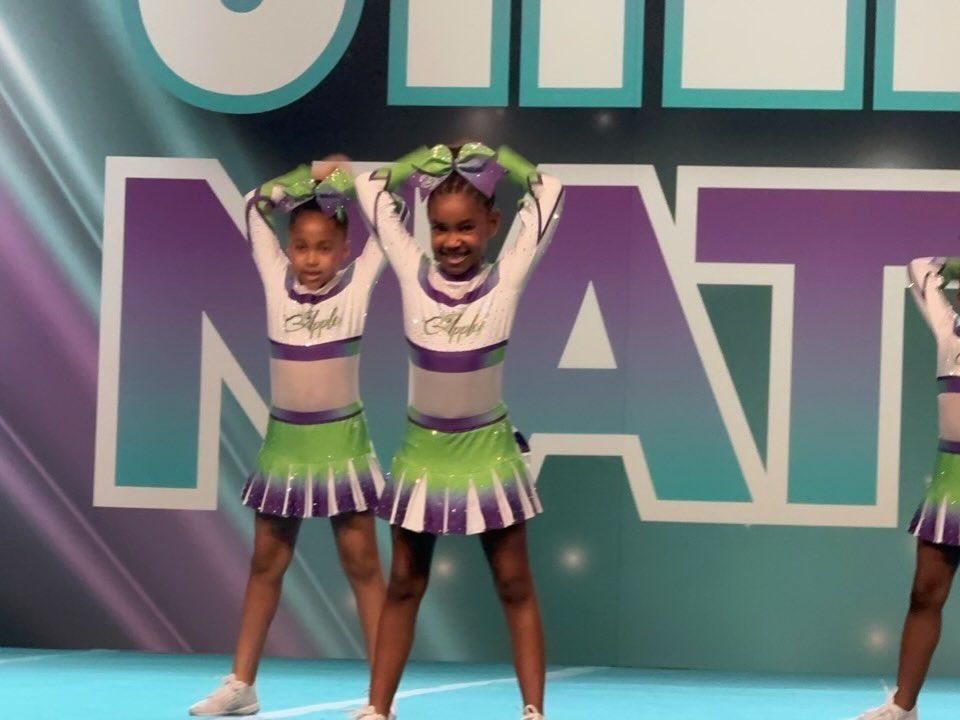 Two young cheerleaders in purple and green uniforms pose with arms raised on a stage.