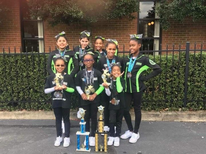 Seven youth holding trophies pose in matching black and green outfits in front of a brick building.