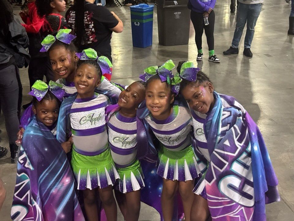 Five smiling cheerleaders in purple, white, and green uniforms pose indoors with matching bows and capes.