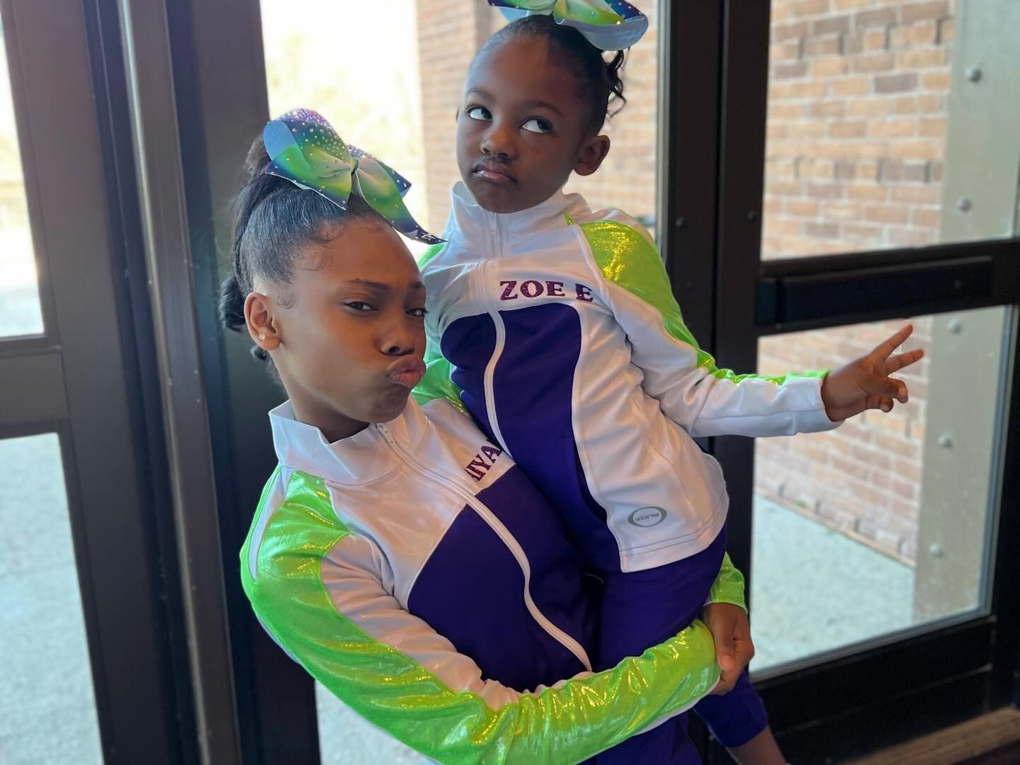 Two cheerleaders in white, purple, and green uniforms posing indoors by a glass door