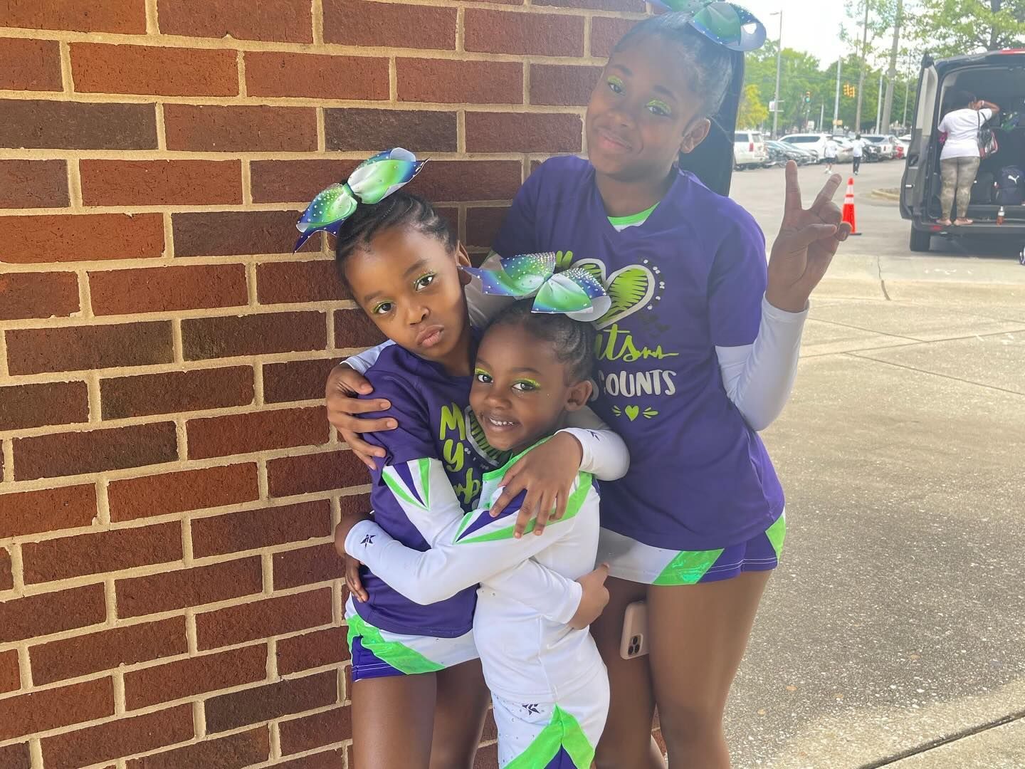 Three children in purple and white outfits posing and smiling by a brick wall outdoors
