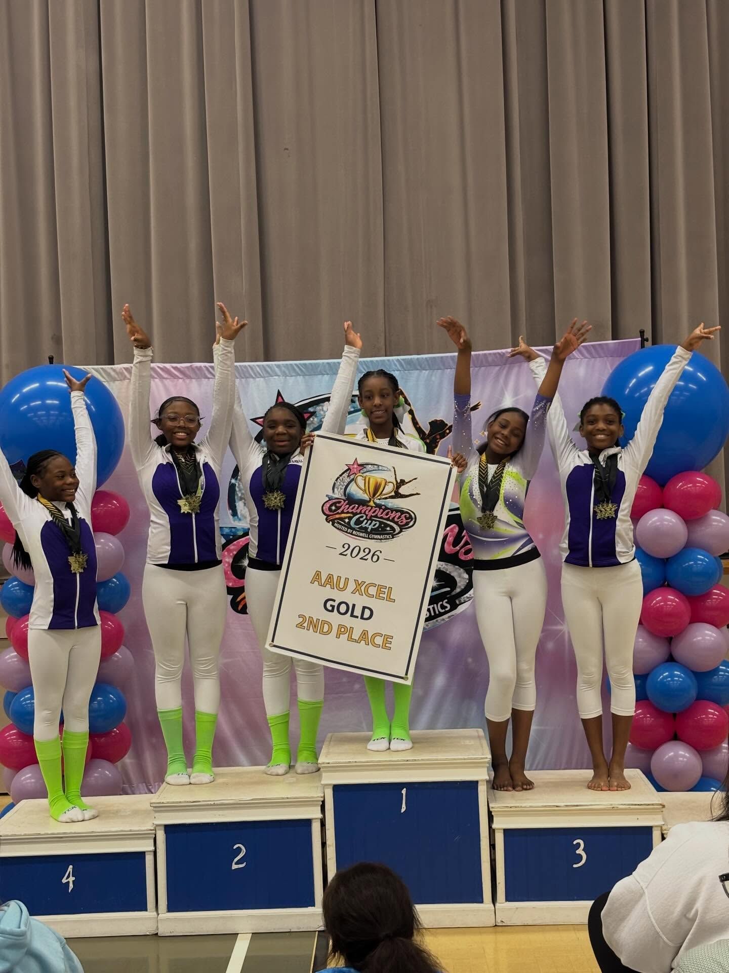 Gymnasts posing on a podium with a sign reading “Angels in the Gym,” holding medals and raising arms.