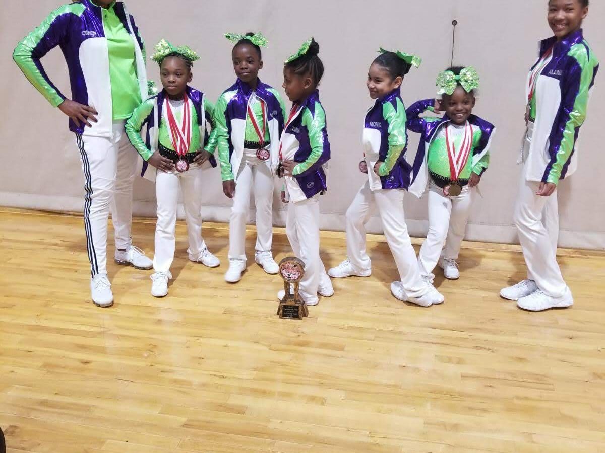 Children in matching purple, green, and white outfits posing on a gym floor with medals and a trophy