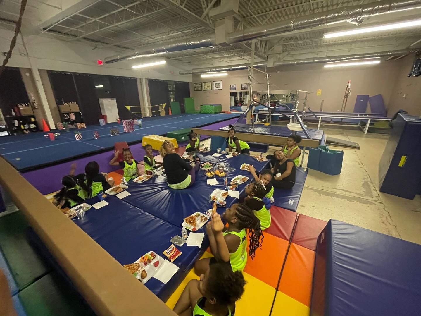 Children doing crafts on mats in a gymnasium, with colorful play equipment and padded floor.
