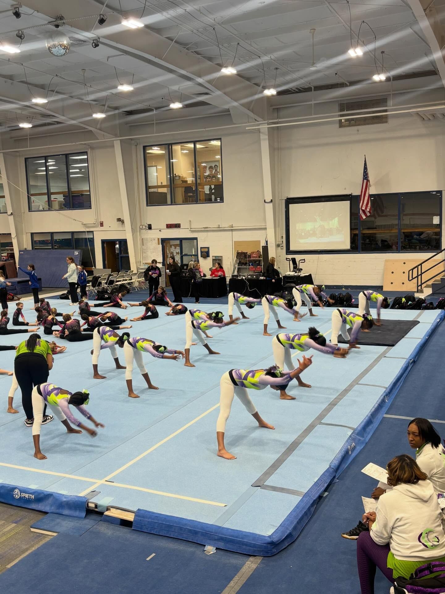 Gymnastics class stretching on blue mats inside a large indoor gymnasium.