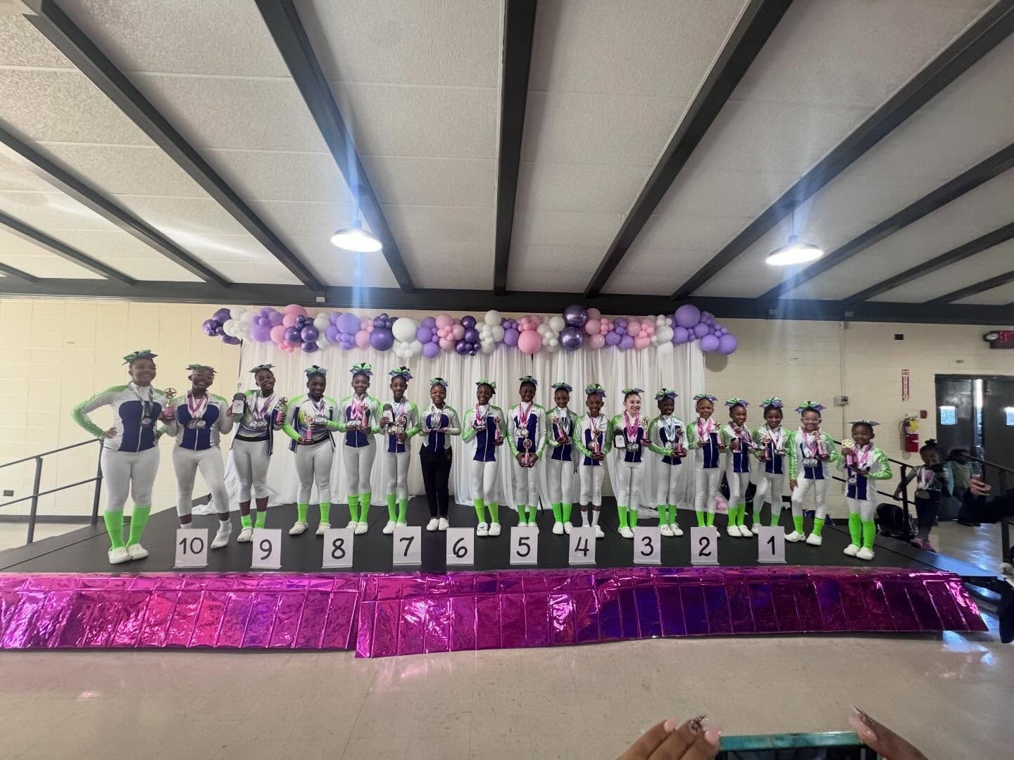 Dance team posing on a decorated stage with purple balloon backdrop and white trophies numbered 1–10.