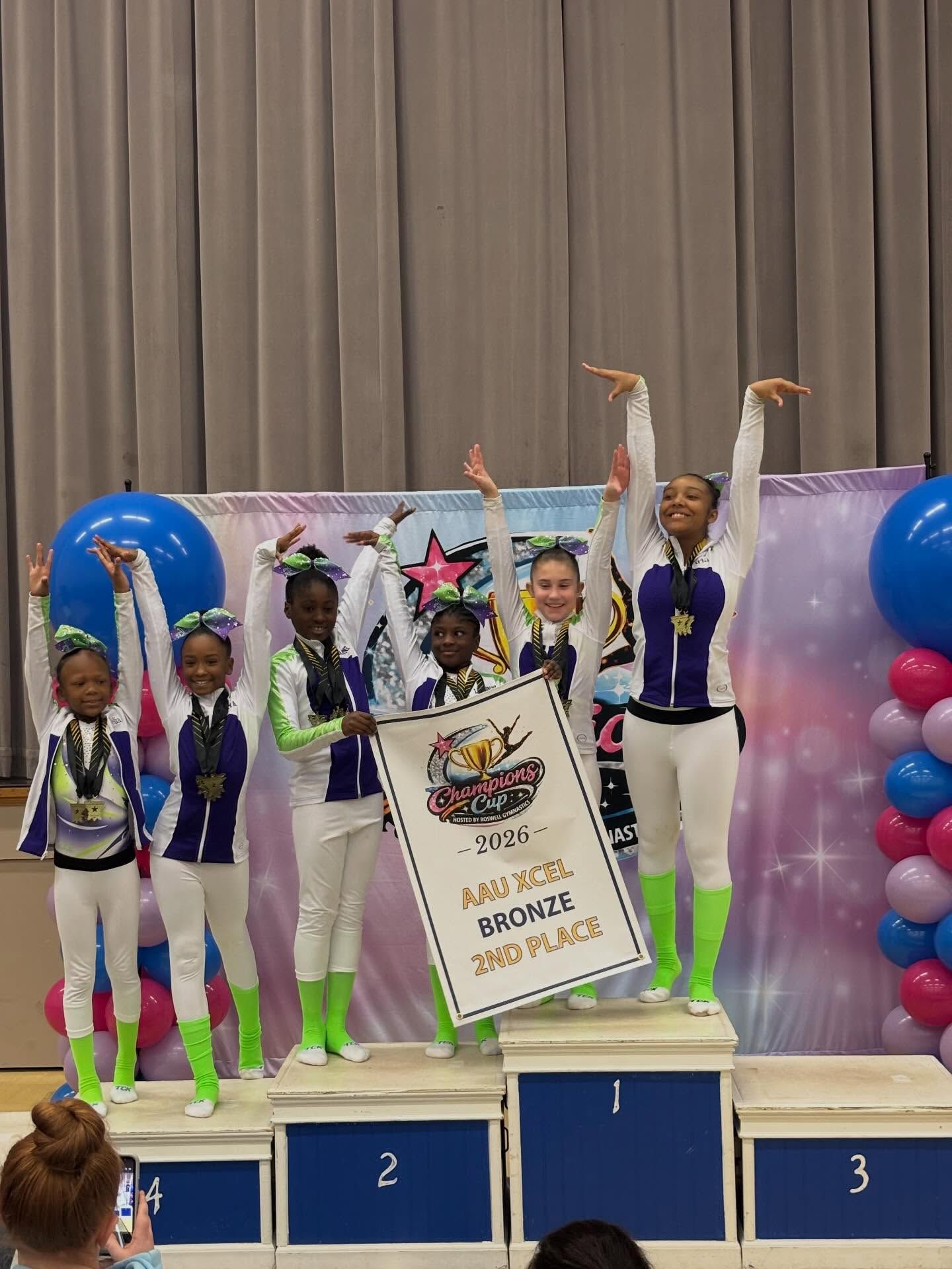 Gymnasts posing on a winners' podium with medals and a large first-place certificate backdrop