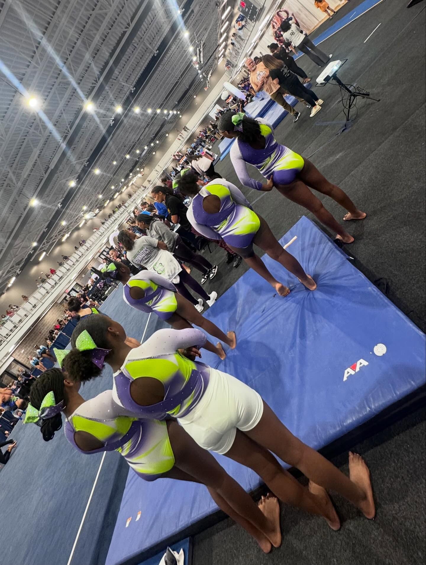 Athletes in purple and yellow uniforms crouch on blue mats in a brightly lit indoor track facility.