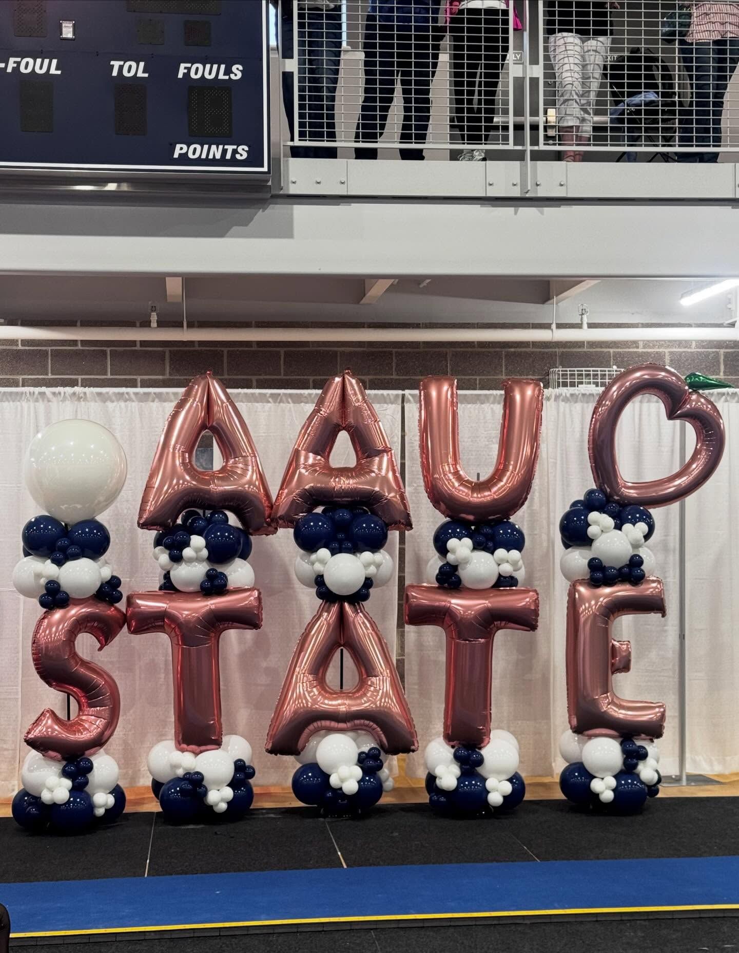 Maroon balloon letters spelling “AUC STATE” with blue, white, and black balloons in a gym.