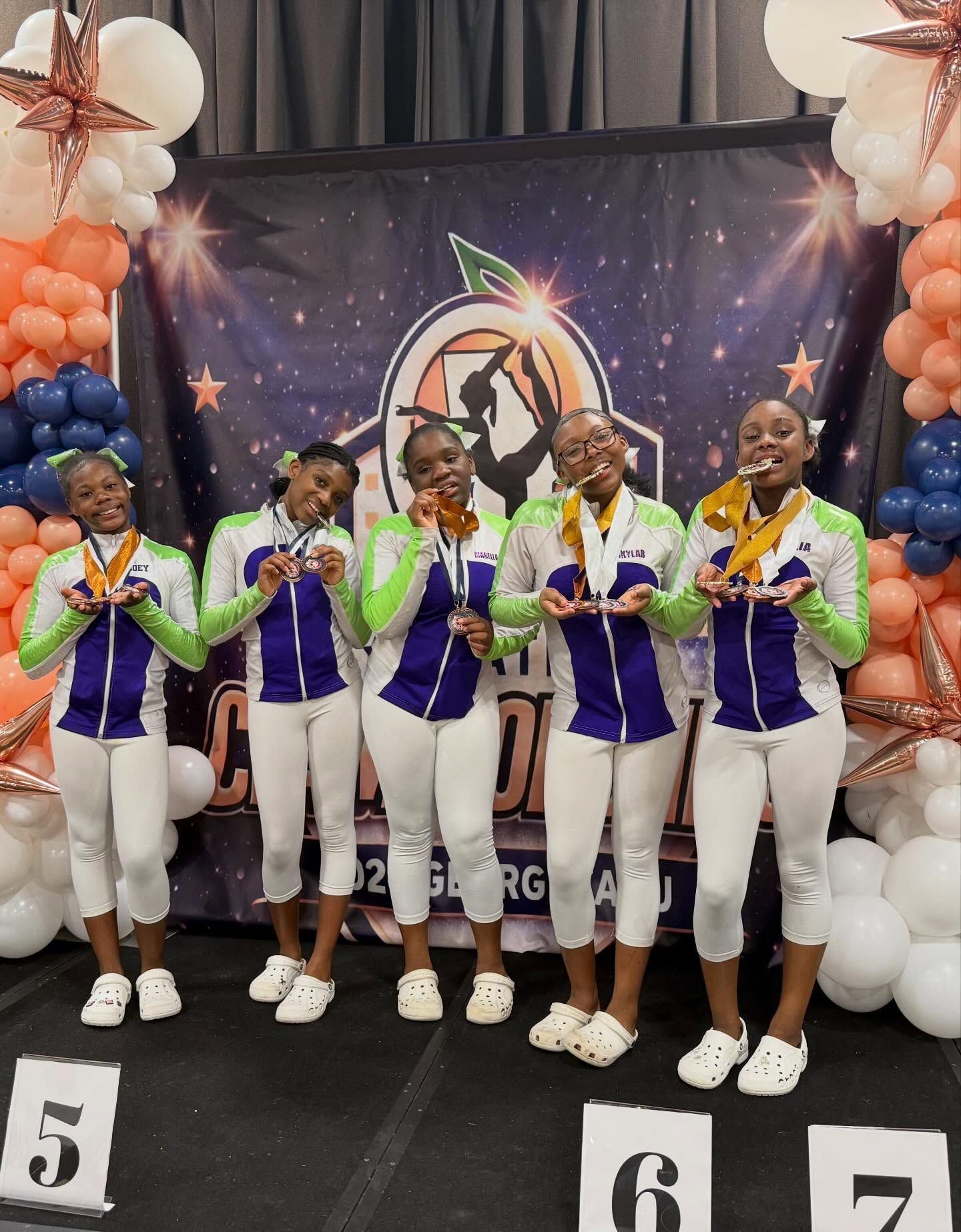 Six dancers in purple and white uniforms posing with medals on a decorated stage with balloons.
