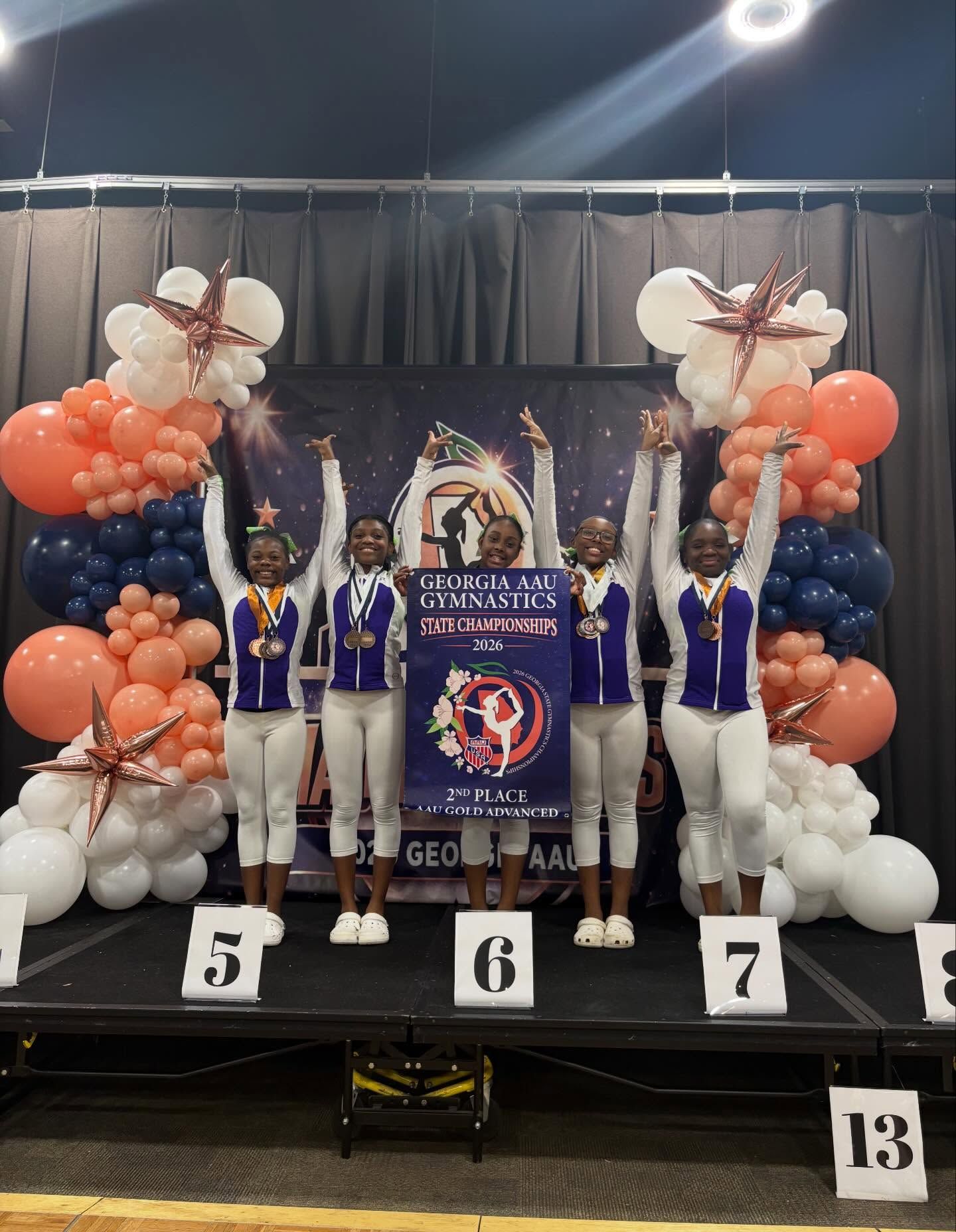 Podium ceremony with four athletes holding a banner amid white and orange balloon decorations.