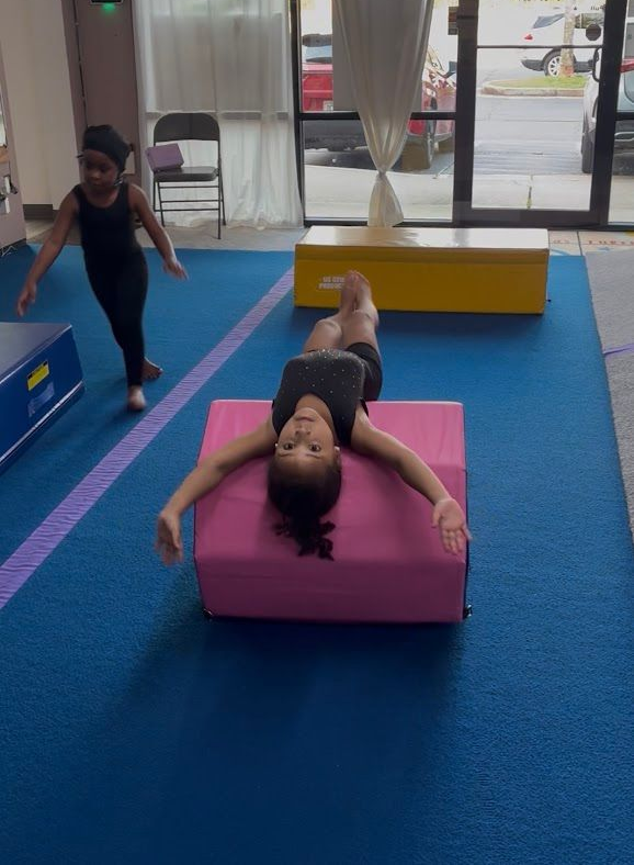Gymnasts in purple and yellow leotards stretching on a blue mat in a gymnasium