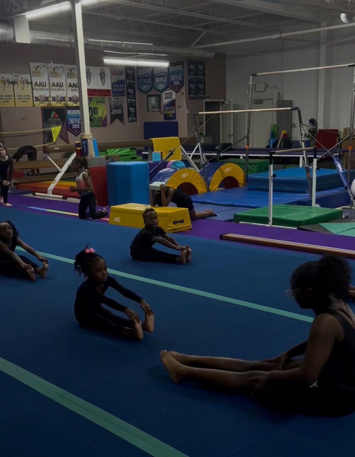 Gymnastics class stretching on blue mats in a large indoor gym.