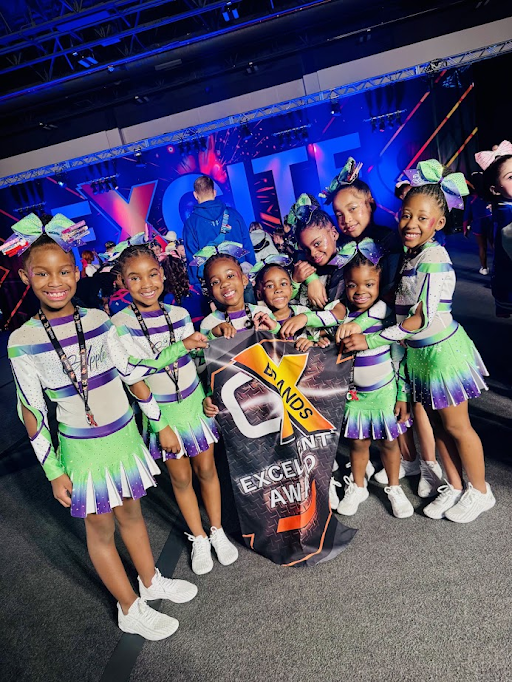 Gymnasts posing on a podium with a sign reading “Angels in the Gym,” holding medals and raising arms.