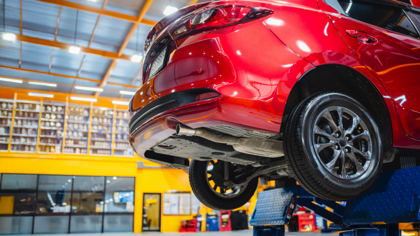 Red car raised on a lift in an auto repair shop with shelves and tools in the background.