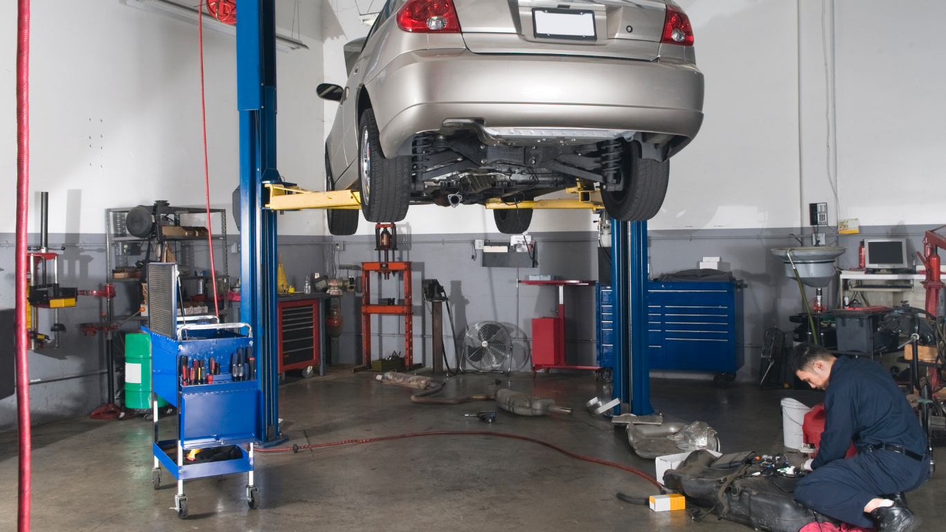 Car lifted on a blue hydraulic hoist in an auto repair shop; mechanic kneels nearby.