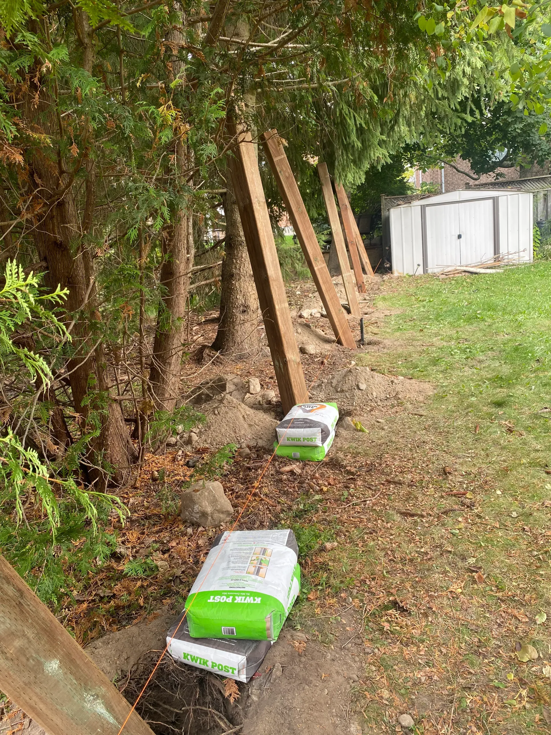 Wooden fence posts being set up with bags of concrete. Backyard setting with grass and trees.