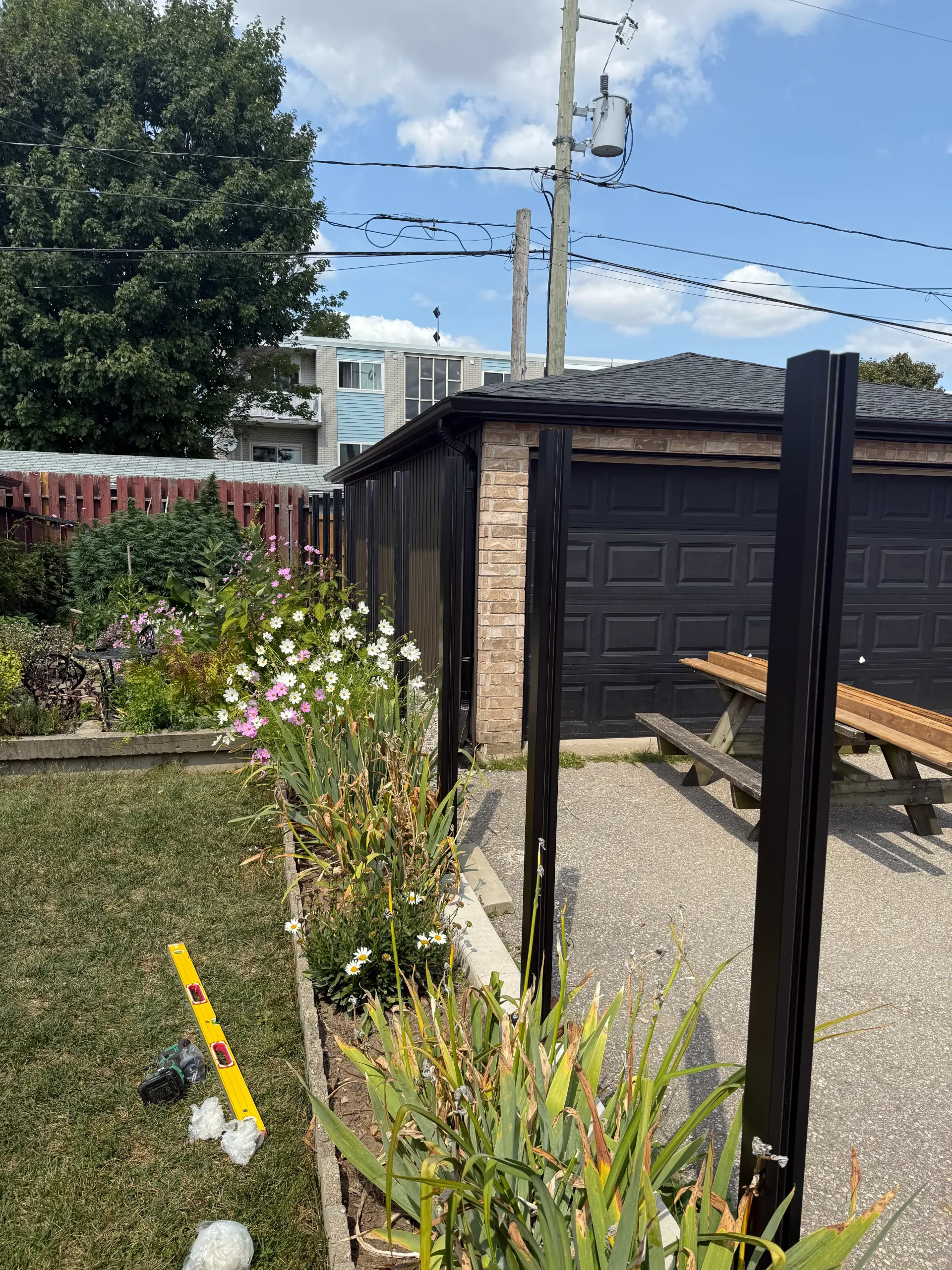 Backyard with a flower bed, gravel path, and garage. Dark vertical posts stand in the foreground.