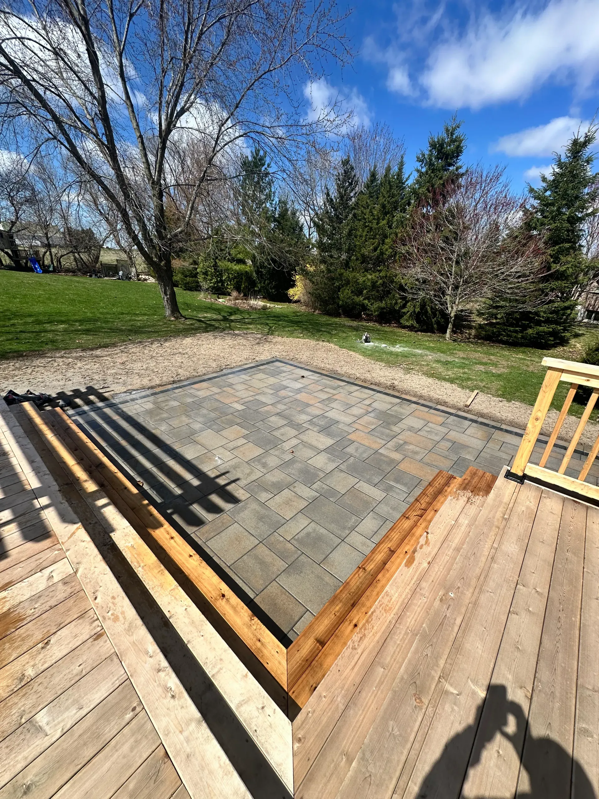 Wooden deck with steps leading to a paved patio in a grassy backyard, blue sky.