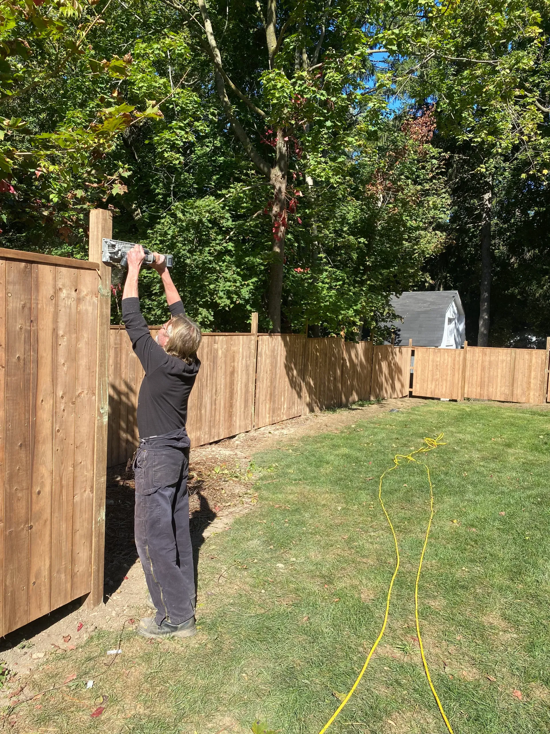 Person using a power tool to work on a wooden fence outdoors.