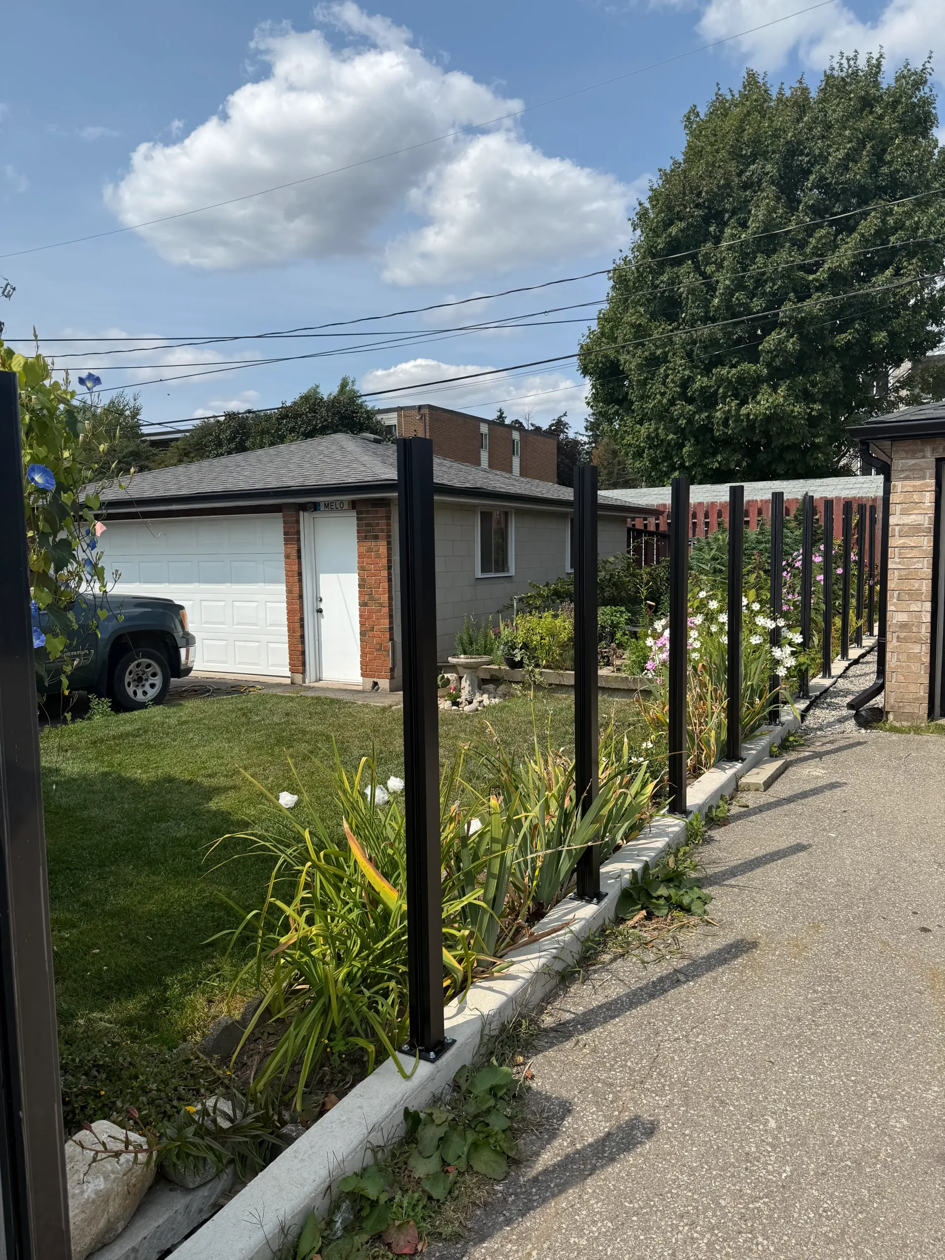 Black metal fence, overgrown garden, gray driveway, white garage with open door, blue sky, and a cloudy day.