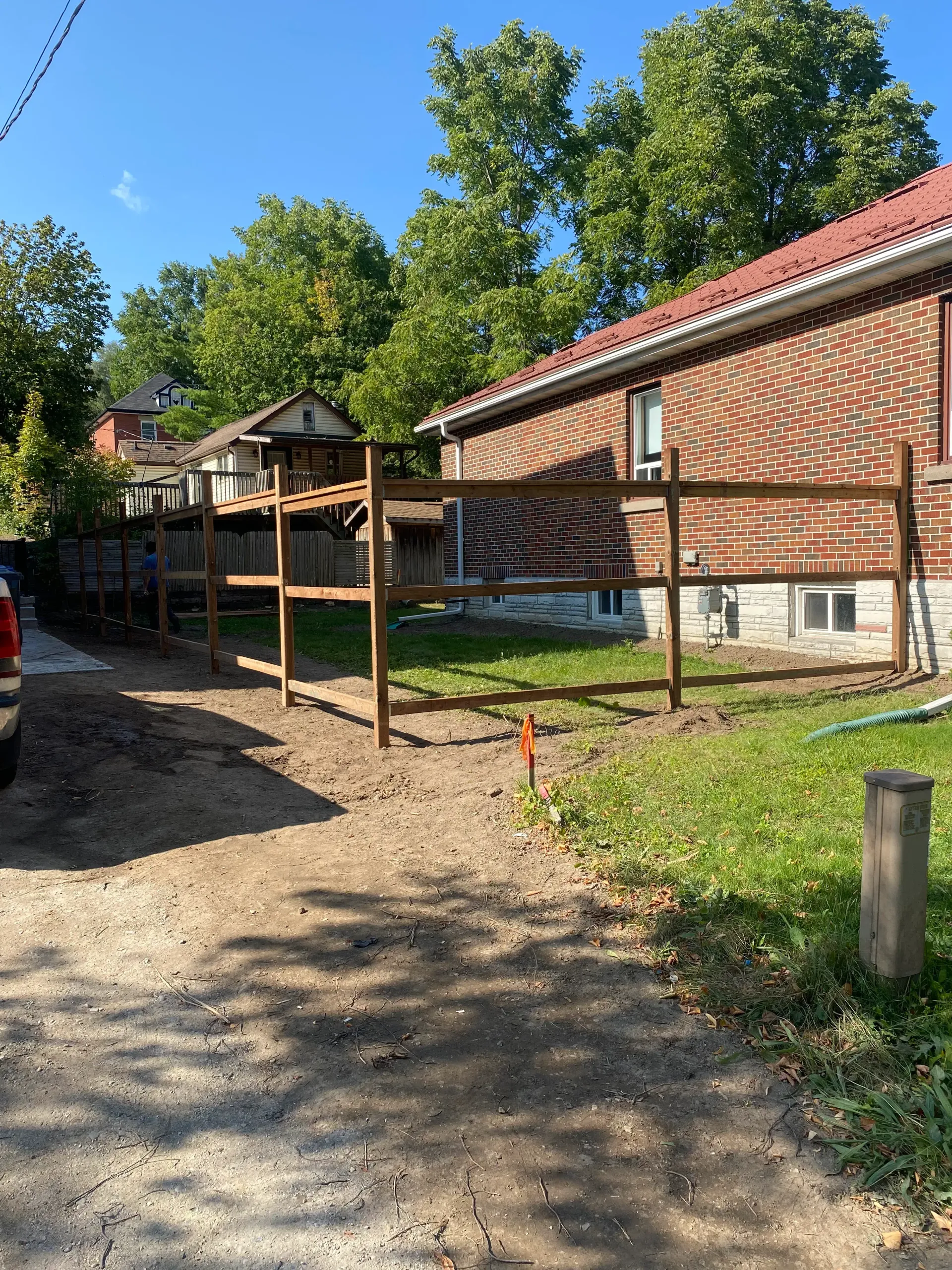 Newly constructed wooden fence next to a brick building. Gravel driveway in the foreground. Green trees and blue sky.