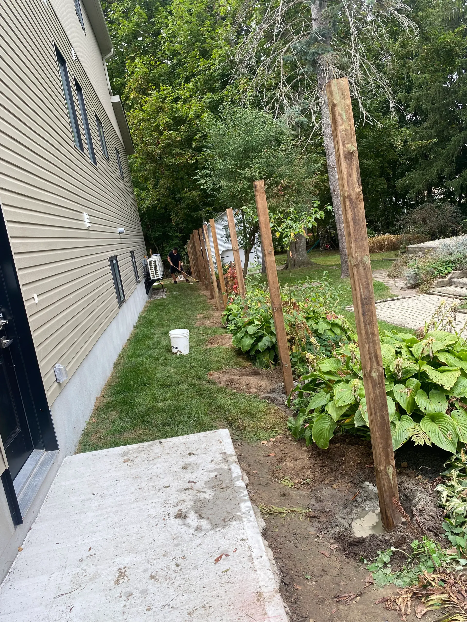 Wooden fence posts along a house with new siding and a small concrete walkway. Green plants in the yard.