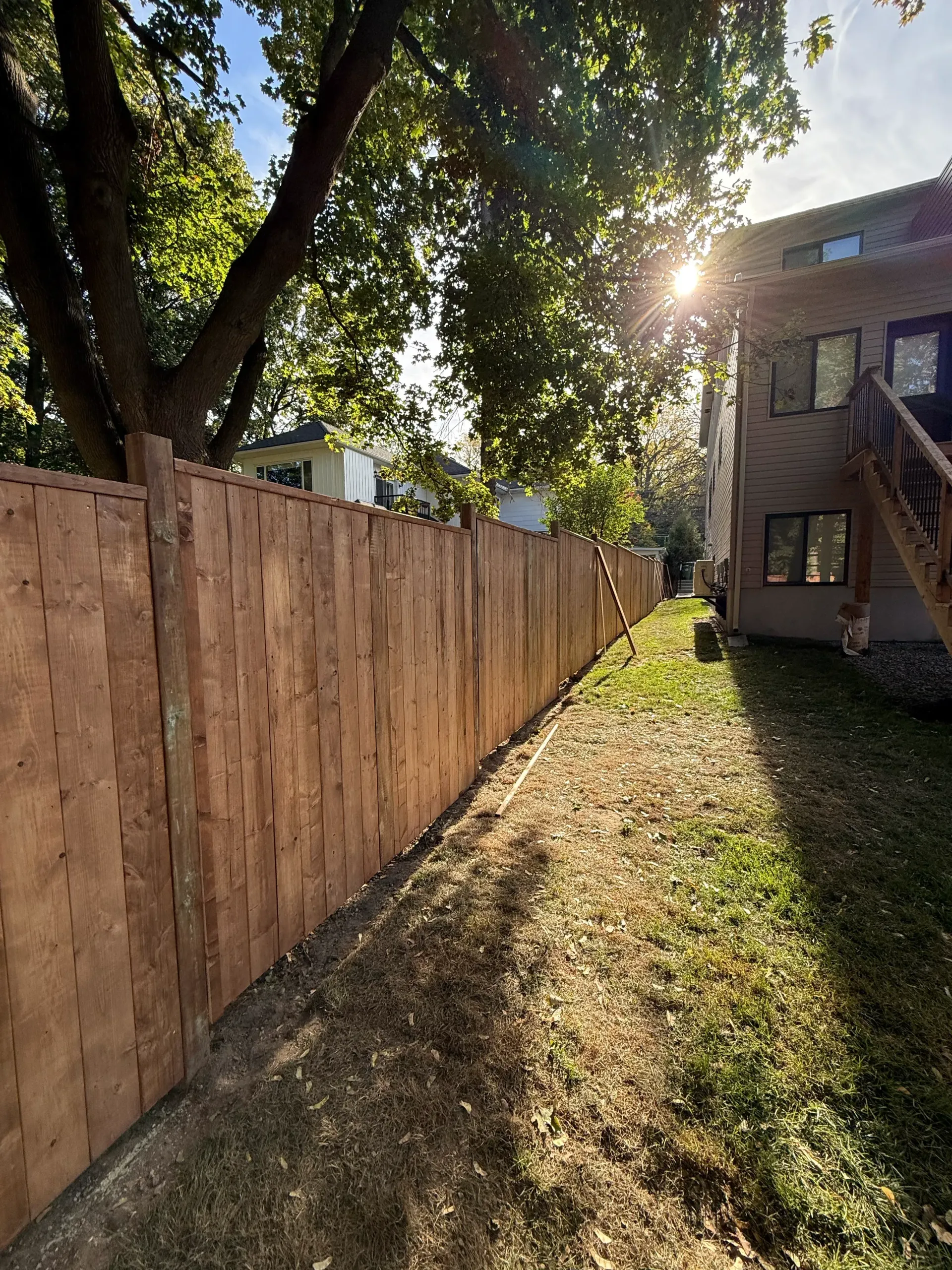 Wooden fence along a grassy path with a building and tree in the background, sun shining.