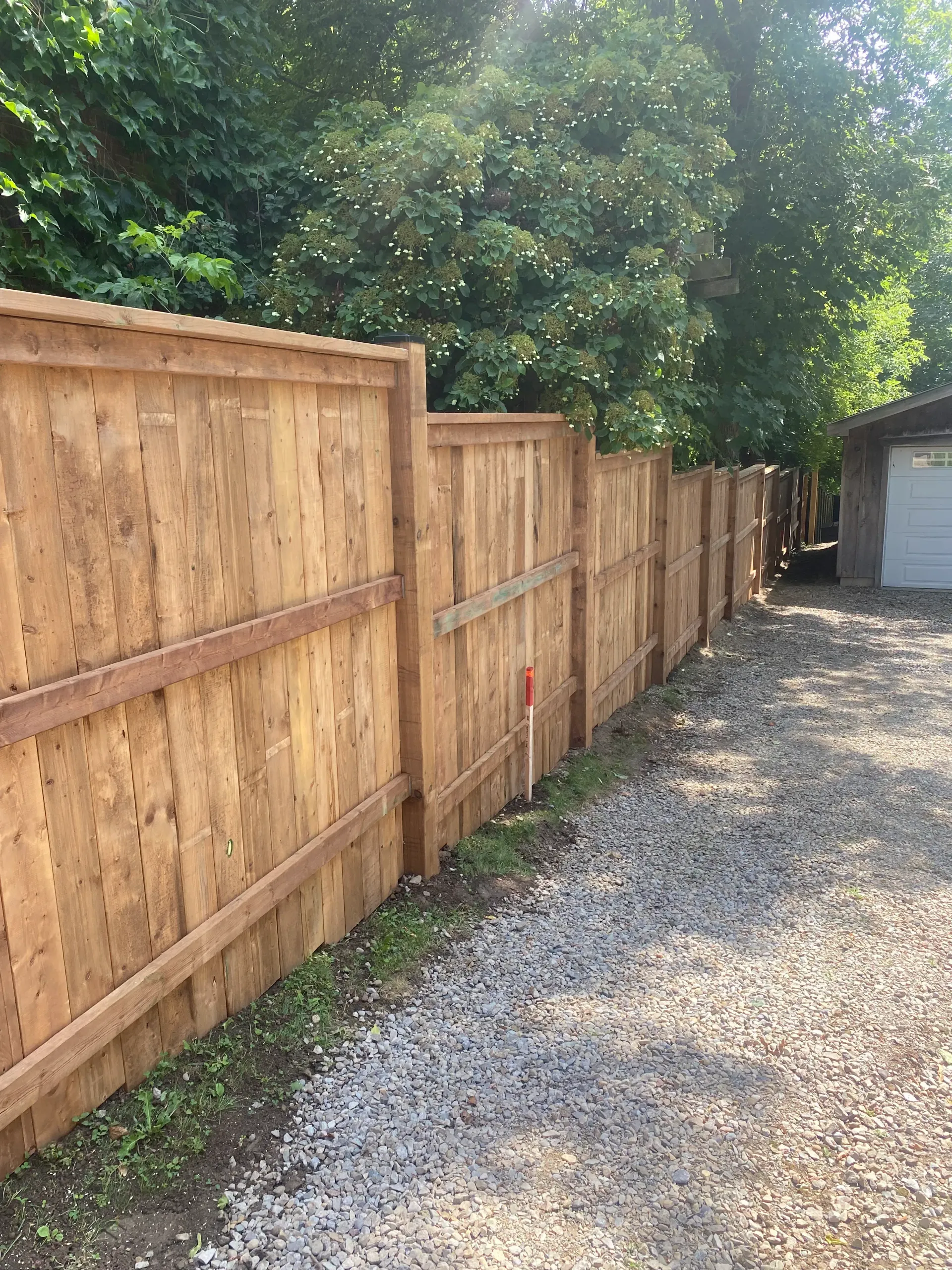 Wooden fence along a gravel driveway; trees in the background, a garage is visible.