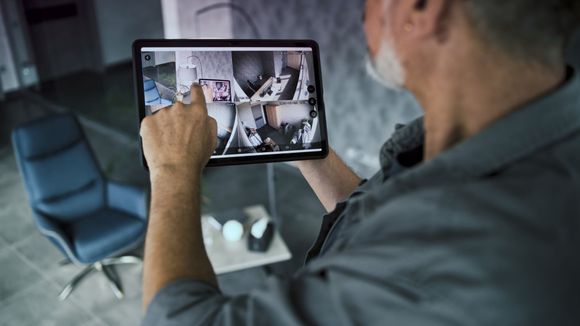 Person viewing a home security camera feed on a tablet, showing several indoor rooms.