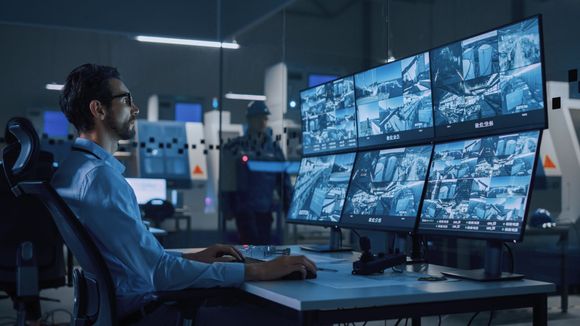 Person at a desk monitoring multiple security camera screens in a dimly lit control room